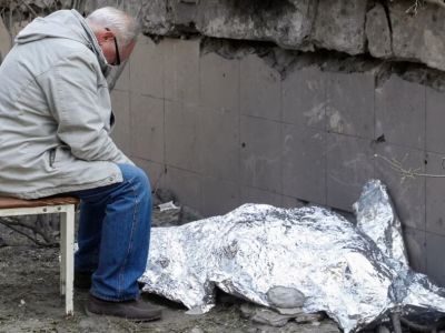 Дедушка сидит рядом с накрытым телом 9-летней внучки возле поликлиники, в которой находится бомбоубежище. Фото: